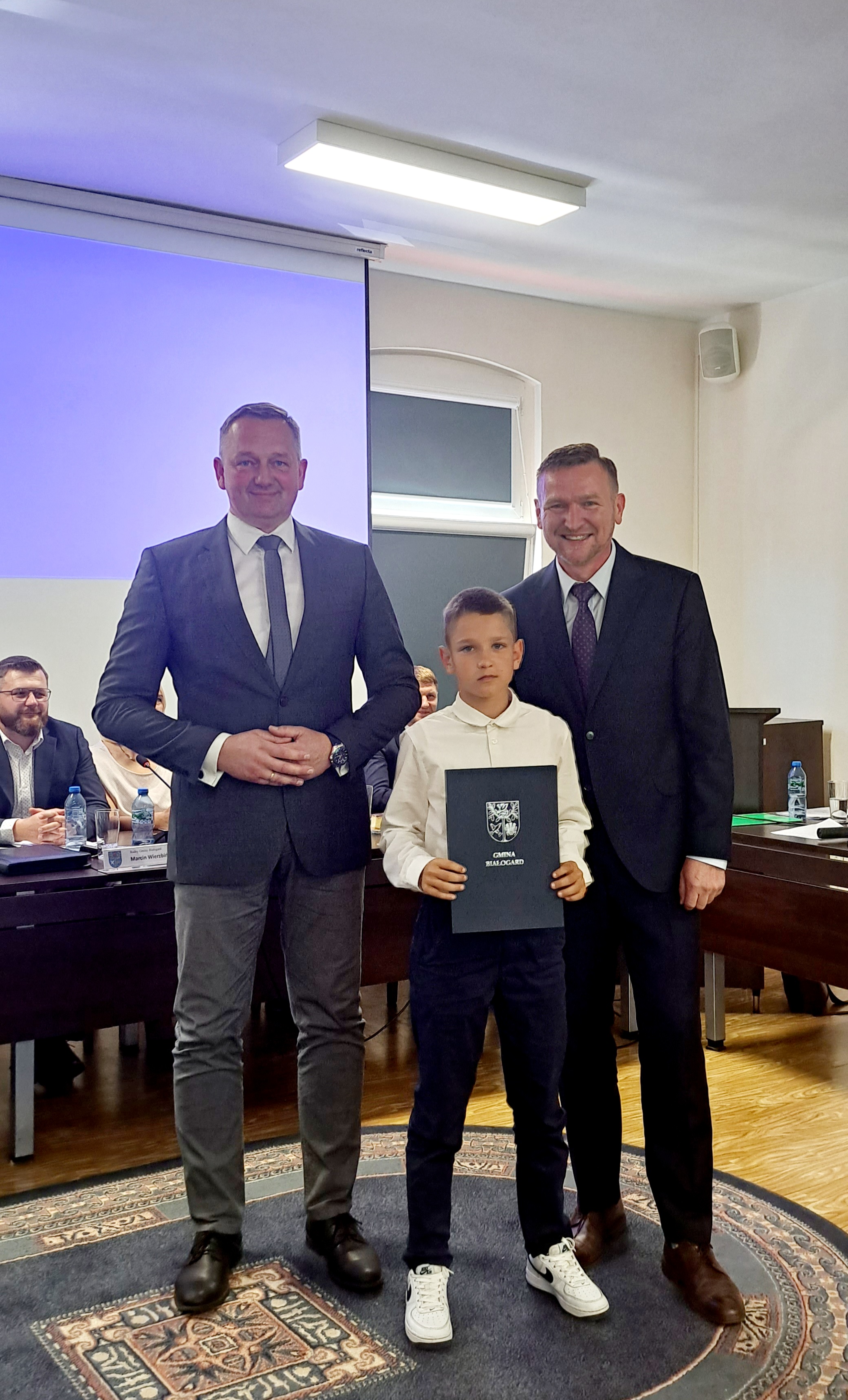 Sala posiedzeń Rady Gminy Białogard. Widoczny z lewej strony Radny siedzący przy stole. W środku trzej panowie. Z lewej Wójt Gminy Białogard Jacek Smoliński, z prawej Przewodniczący Rady Gminy Marcin Pender, w środku nagrodzony sportowiec Ksawery Gajda, trzymający w rękach okładkę z herbem Gminy Białogard.