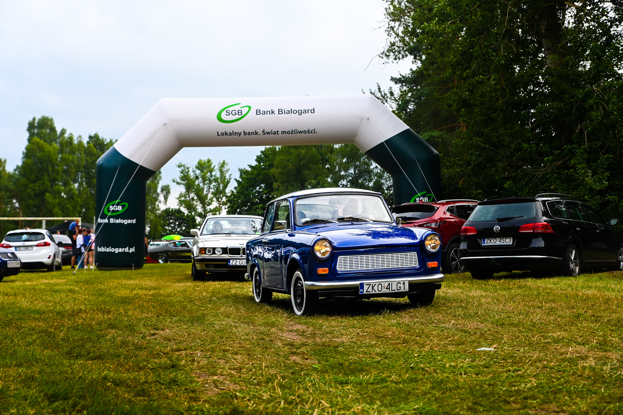 Na zdjęciu widoczny niebieski zabytkowy pojazd marki trabant, w tle różne inne pojazdy zabytkowe oraz balon sponsora Bank Spółdzielczy Białogard