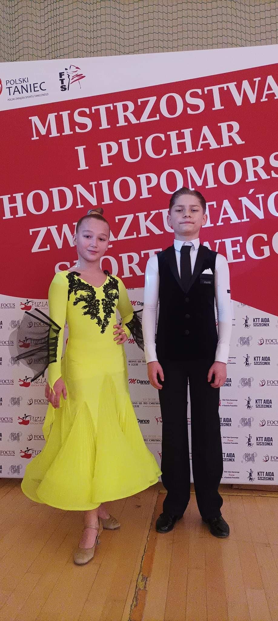 Na zdjęciu Aleksander i Kaja. Kaja ubrana w długą neonową sukienkę w odcieniu żółci z czarnymi dodatkami. Aleksander ubrany w białą koszulę z czarnym krawatem, w czarne spodnie i welurową czarną kamizelkę. Dumnie się prezentują. Na tle widać napis Mistrzostwa i Puchar Zachodniopomorskiego Związku Tańca Sportowego.