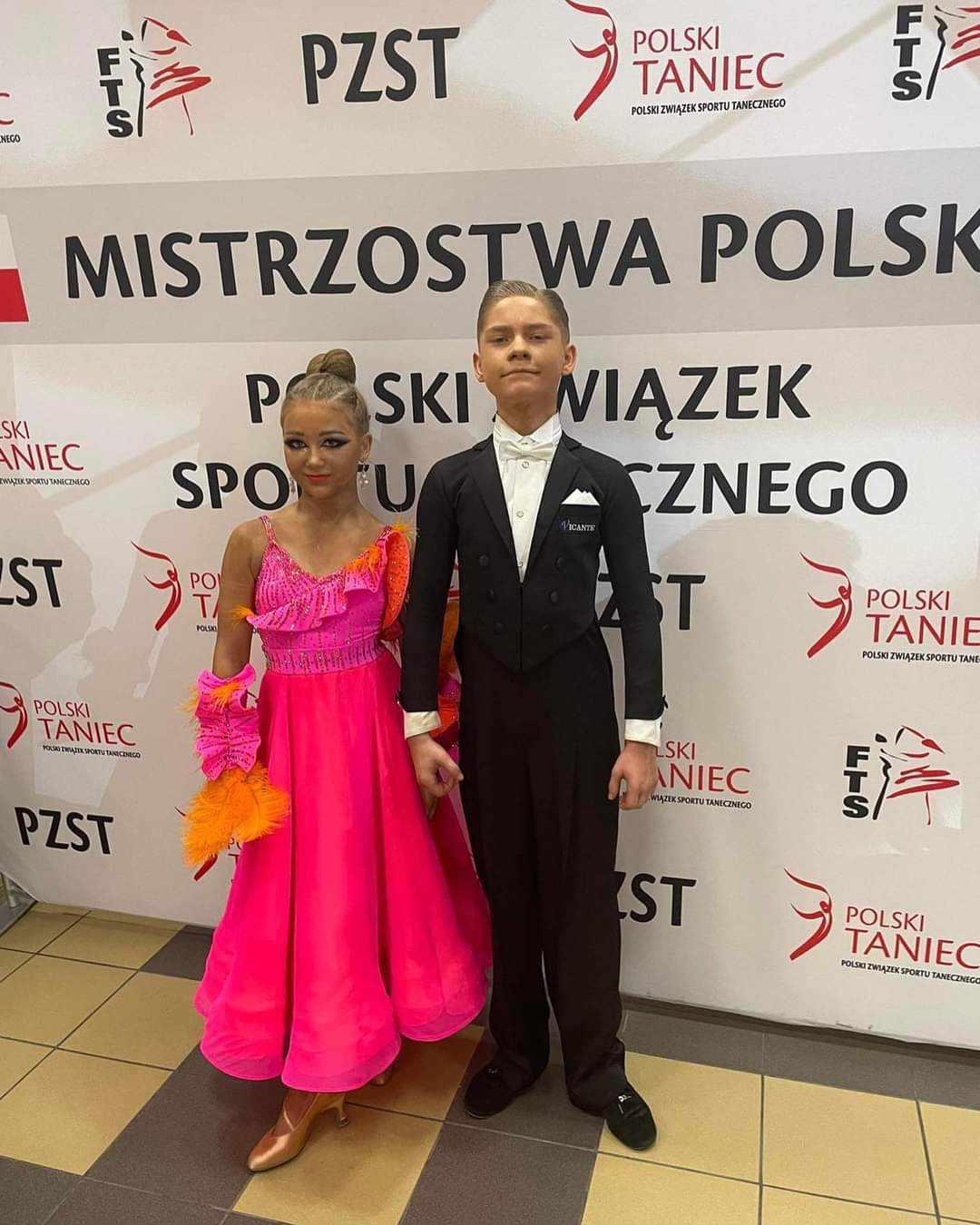 Na tle baneru Mistrzostwa Polski stoi Aleksander Dondera z Nosówka i Kaja Drętkiewicz z Mścic. Aleksander ubrany jest w czarny frak oraz białą koszulę z długim rękawem. Na szyi ma założoną białą muchę. Kaja natomiast ma na sobie długą różowo - pomarańczową sukienkę z doszytymi do niej pomarańczowymi piórami. Uszy zdobią połyskujące kolczyki. Zawodnicy są profesjonalnie uczesani – Aleksander ma zaczesane włosy do góry, natomiast Kaja ma zrobiony kok. Uśmiechają się.