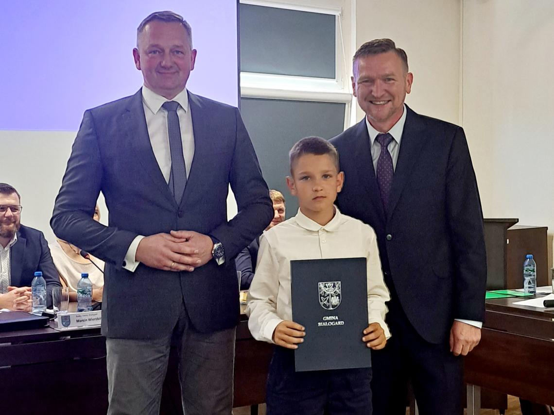 Sala posiedzeń Rady Gminy Białogard. Widoczny z lewej strony Radny siedzący przy stole. W środku trzej panowie. Z lewej Wójt Gminy Białogard Jacek Smoliński, z prawej Przewodniczący Rady Gminy Marcin Pender, w środku nagrodzony sportowiec Ksawery Gajda, trzymający w rękach okładkę z herbem Gminy Białogard.