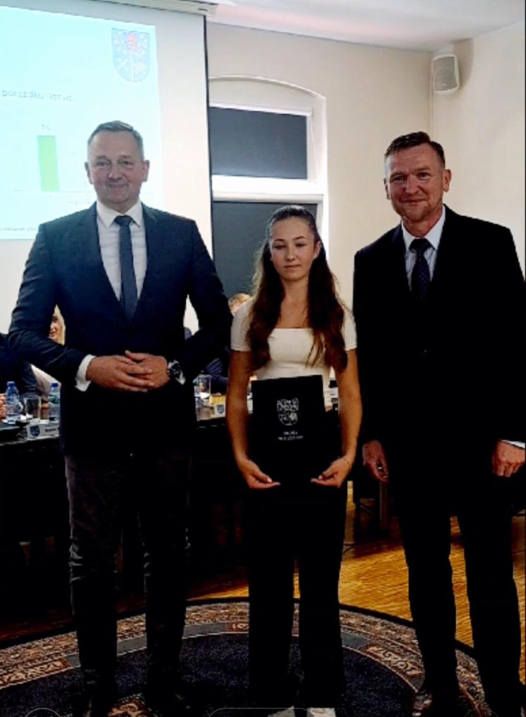 Sala posiedzeń Rady Gminy Białogard. W środku trzy osoby. Z lewej Wójt Gminy Białogard Jacek Smoliński, z prawej Przewodniczący Rady Gminy Marcin Pender,  w środku nagrodzona lekkoatletka Nokola  Żórawska, trzymająca w rękach okładkę z herbem Gminy Białogard.